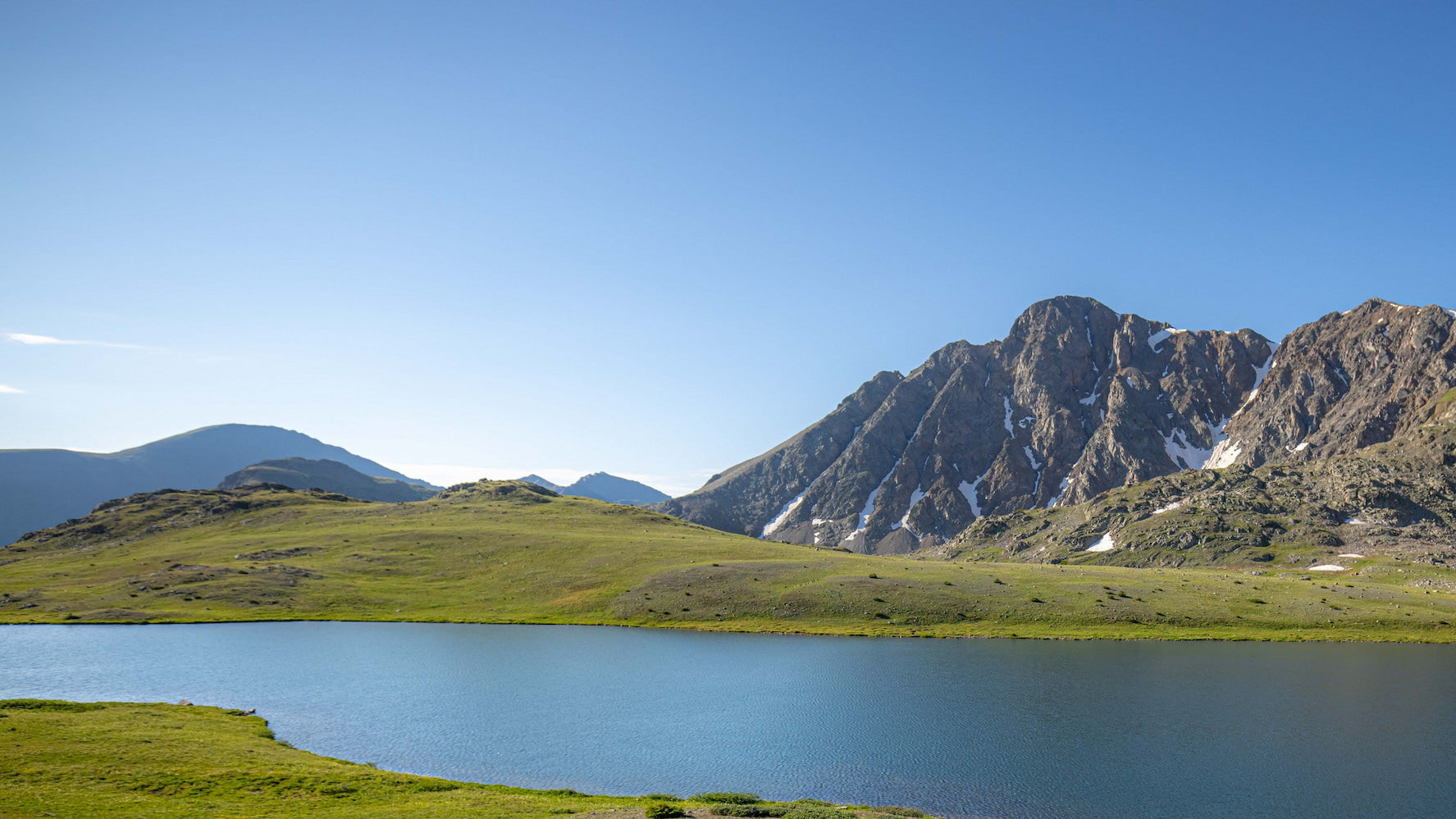 A clear blue lake sits in the foreground with green grassy hills and rocky mountains in the background under a bright blue sky.