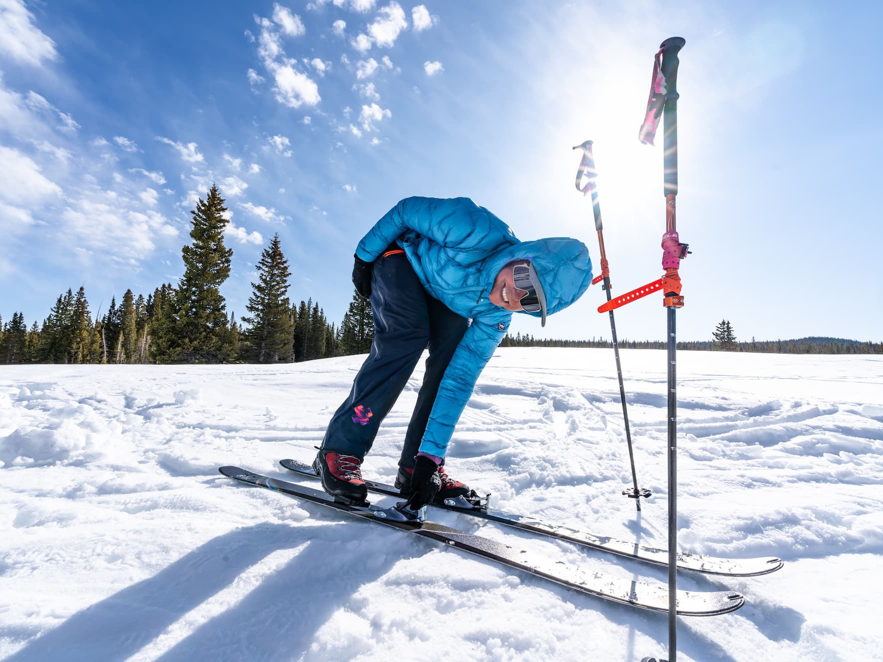 A person in a blue jacket and ski gear bends down to adjust a ski binding on a snowy slope, with bright sunlight and ski poles standing upright nearby. Pine trees and a blue sky with clouds are in the background.