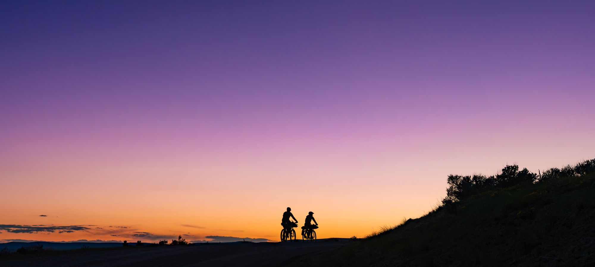 Two people ride bicycles along a hill at sunset, silhouetted against a vibrant sky fading from orange near the horizon to deep purple above, with trees and bushes visible on the right.