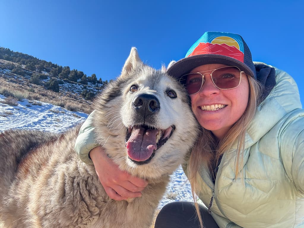 A woman wearing sunglasses and a colorful cap smiles and hugs a large, happy, fluffy dog outdoors on a sunny, snowy day with hills and blue sky in the background.