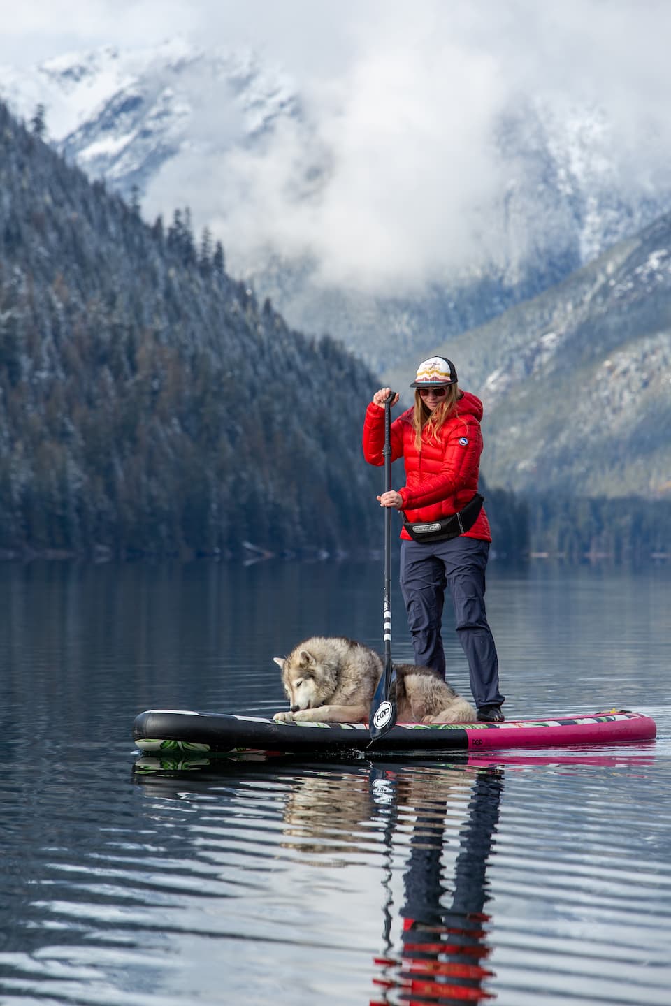 A woman in outdoor gear stands on a paddleboard with a large, wolf-like dog lying beside her, surrounded by calm lake water and snow-capped mountains in the background.