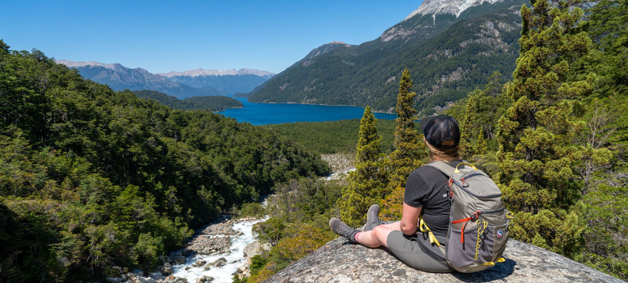 A person with a backpack sits on a rock, overlooking a lush green valley with a river, forest, and a blue lake surrounded by mountains under a clear sky.