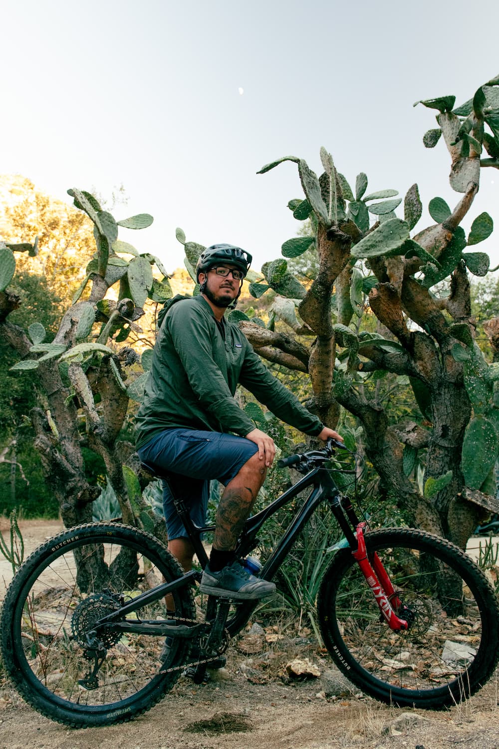 A man wearing a helmet and glasses sits on a mountain bike on a dirt path, surrounded by large cactus plants, with sunlight filtering through the background.