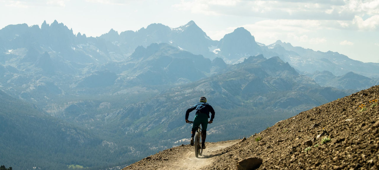 A mountain biker rides down a dirt trail on a hillside, surrounded by sprawling mountain ranges under a partly cloudy sky. The scene is rugged and expansive, with distant peaks in the background.