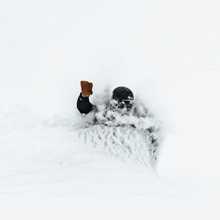 A skier wearing a helmet and brown glove is almost completely submerged in deep powdery snow, with only their head and raised arm visible above the white surface.