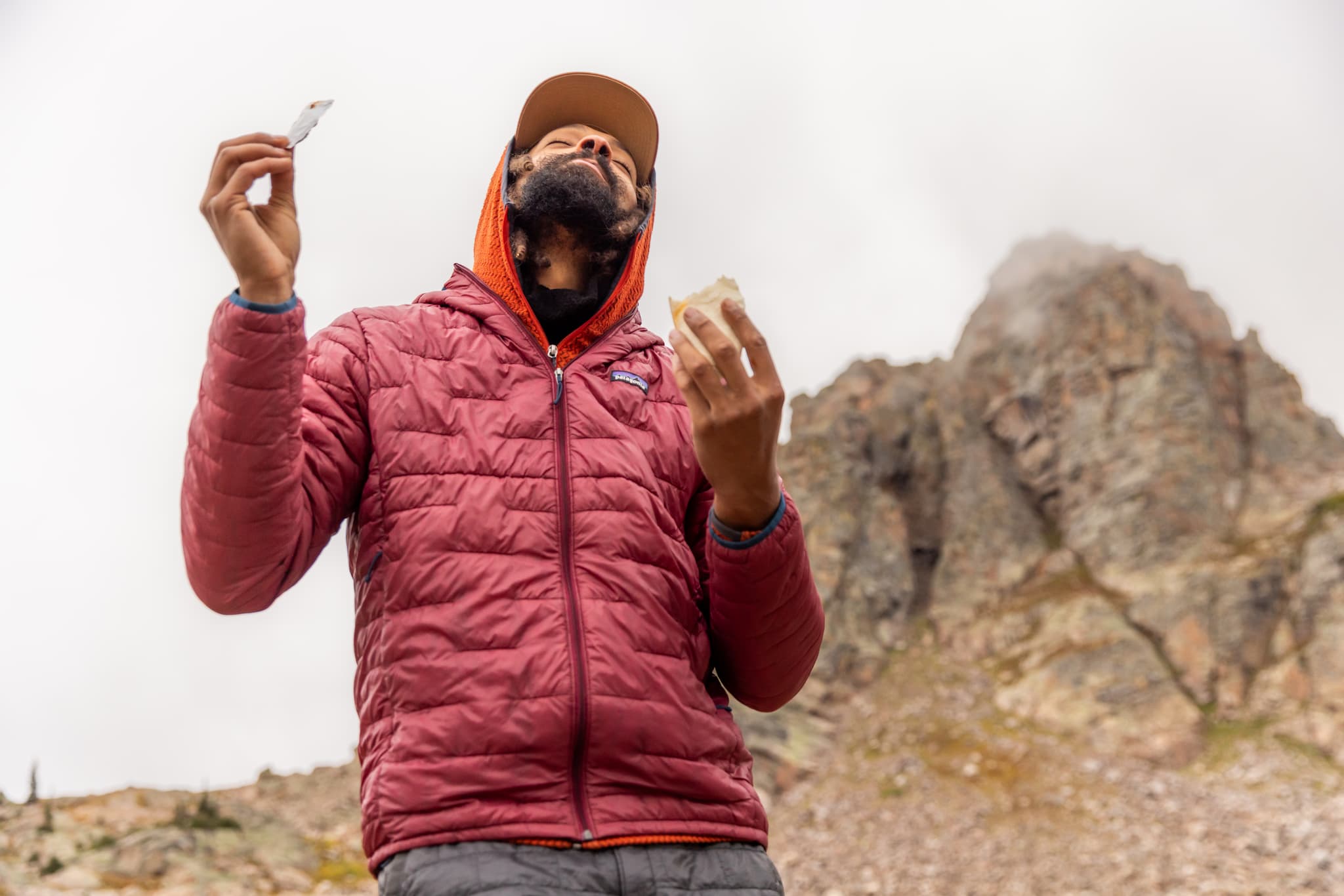 A person wearing a red jacket and orange hood stands outdoors in front of a rocky mountain, holding food in both hands and looking upwards, with a cloudy sky overhead.