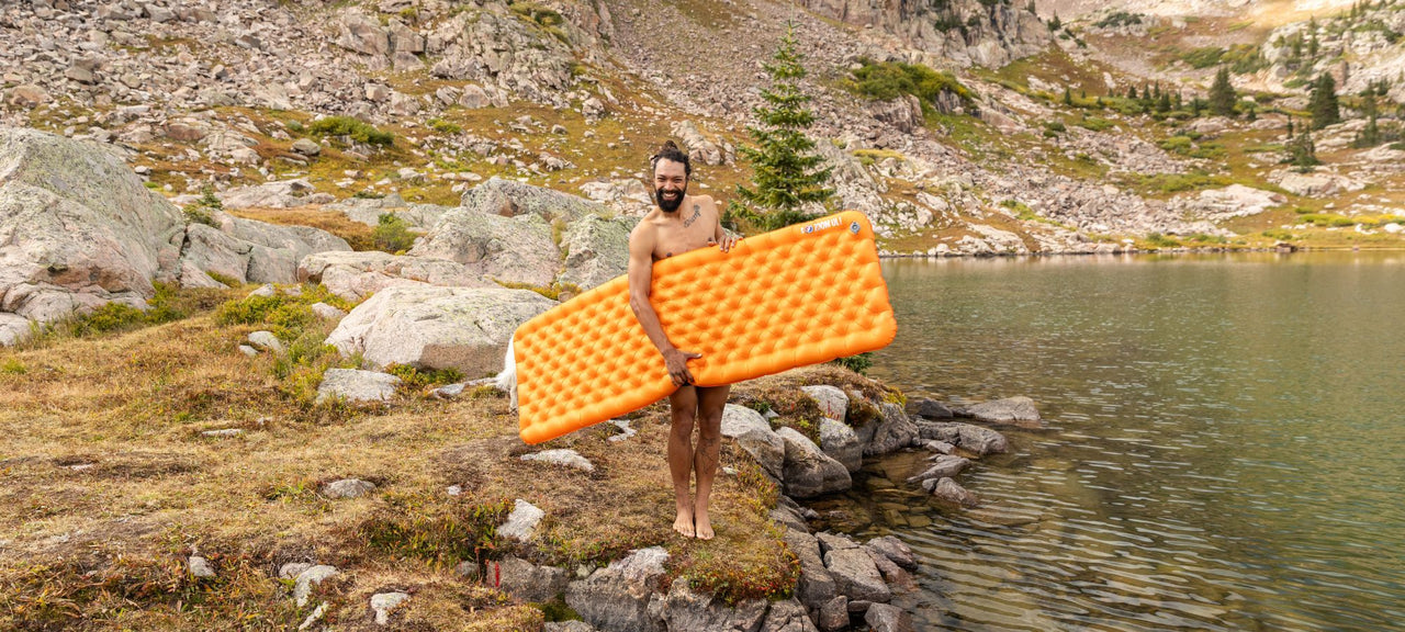 A man stands barefoot by a mountain lake, holding an orange inflatable mattress and smiling. Rocky terrain and greenery surround the water, with mountains in the background.