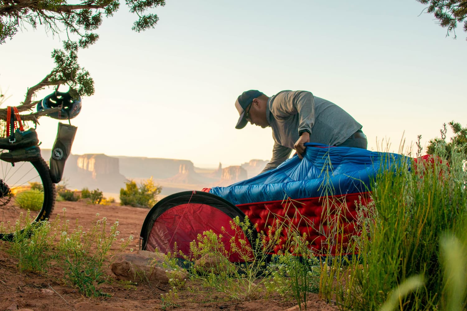 A person in a cap sets up a bright red and blue sleeping bag next to a small tent in a desert campsite with green shrubs and distant mesas at sunrise; a bicycle is parked nearby.