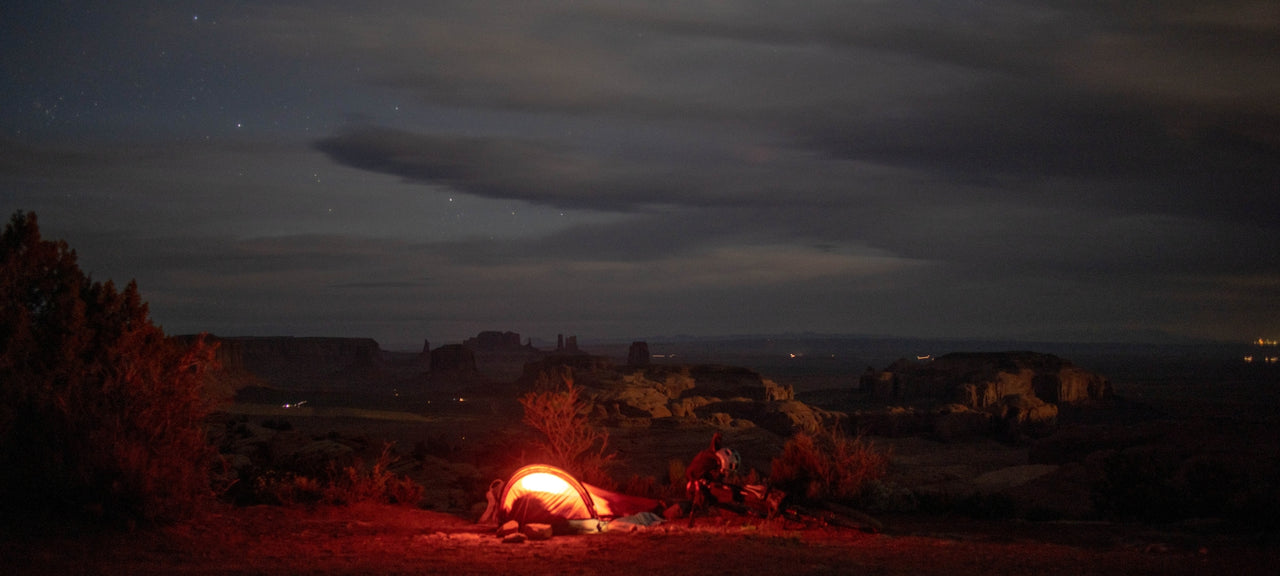 A glowing tent is set up in a vast, rocky desert landscape at night under a cloudy, starry sky, with distant mesas silhouetted against the horizon.