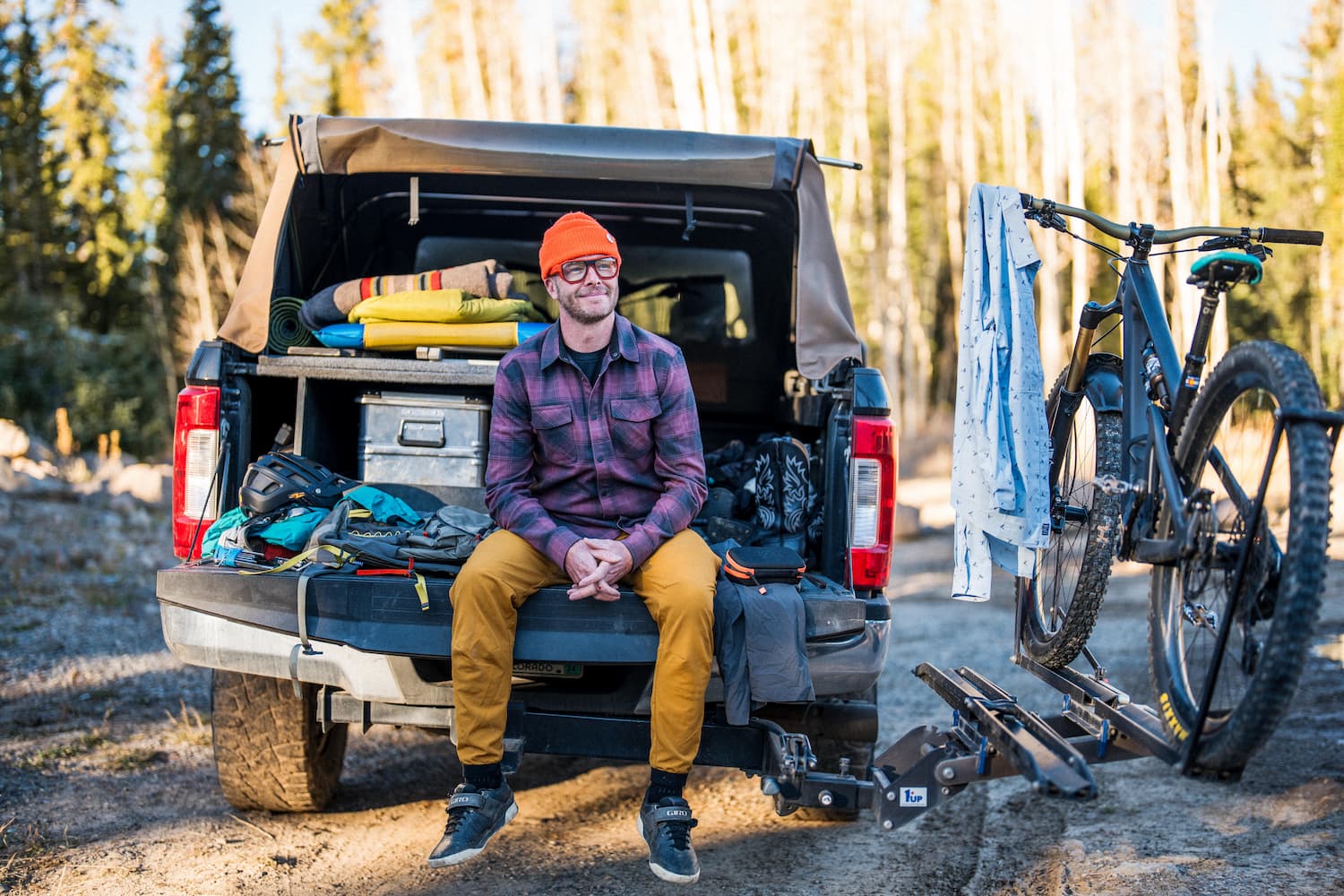 A man wearing an orange beanie and glasses sits on the tailgate of a pickup truck, surrounded by outdoor gear and a bike mounted on the back, with trees and sunlight in the background.