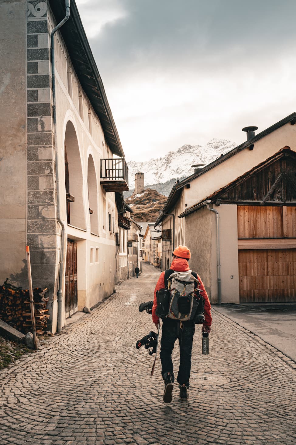 A person wearing a red jacket and backpack walks down a cobblestone street in a mountain village, carrying skis and poles, with snow-capped peaks visible in the distance under a cloudy sky.