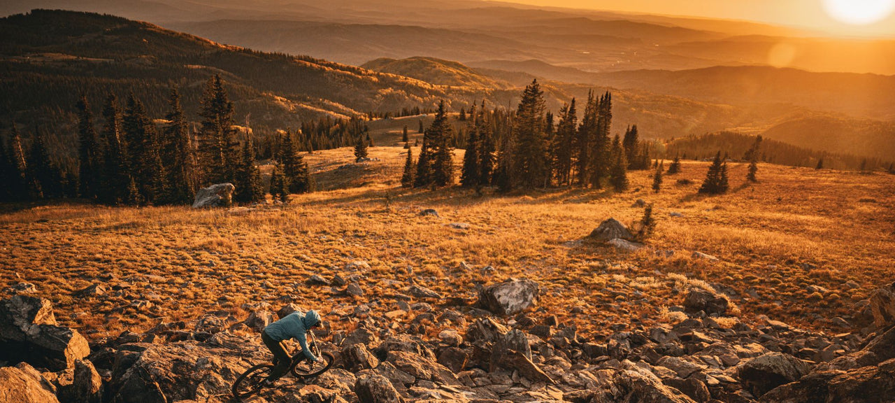 A person rides a mountain bike over rocky terrain at sunset, surrounded by pine trees and rolling hills with golden light illuminating the landscape.