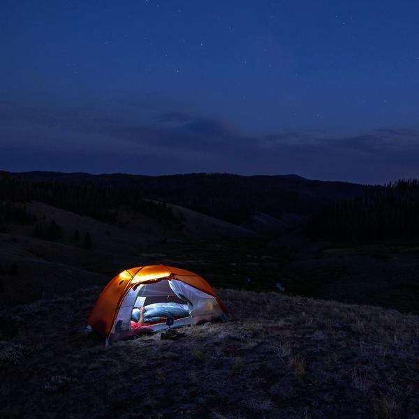 A glowing orange Big Agnes Canada Copper Spur UL2 mtnGLO® tent with integrated lighting stands on a grassy hilltop under a clear, star-filled sky, encircled by rolling dark hills and a distant forest.