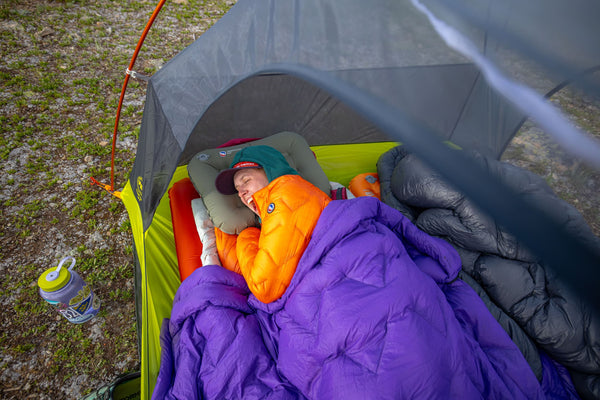 A person wearing an orange jacket and green hat sleeps inside a tent, covered with a purple sleeping bag. An insulated water bottle sits on the ground beside the tent. The ground outside is grassy and rocky.