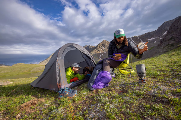 Two people at a mountain campsite; one sits outside the tent cooking with a portable stove, while the other relaxes inside the tent. The sky is partly cloudy, and rugged mountains are in the background.