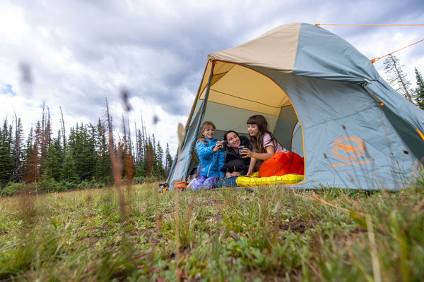Three people sit at the entrance of a tent on a grassy field, looking at a phone together, surrounded by pine trees under a cloudy sky. Camping gear and bright sleeping bags are visible inside the tent.