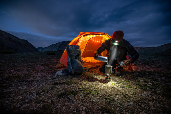 A person sits outside an illuminated orange tent at dusk, surrounded by mountains. They prepare food using a camping stove, with a backpack and gear nearby on the rocky ground.
