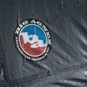 Close-up of the wet, gray grid-patterned Hyperbead fabric on the Pitchpine VST 1.5 by Big Agnes Canada, showing raindrops beading on this ultralight tent’s durable surface and the red, blue, and white logo.