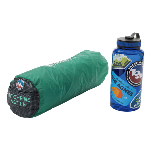 A green cylindrical tent bag labeled Big Agnes Canada Pitchpine VST 1.5 made of Hyperbead fabric rests beside a blue reusable water bottle decorated with outdoor stickers.