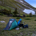 A person in a blue jacket sits on a rock near the Big Agnes Canada Pitchpine VST 1.5 ultralight tent, with DAC Featherlite poles, reading a map in grassy mountains with trees, snow patches, and camping gear beside them.