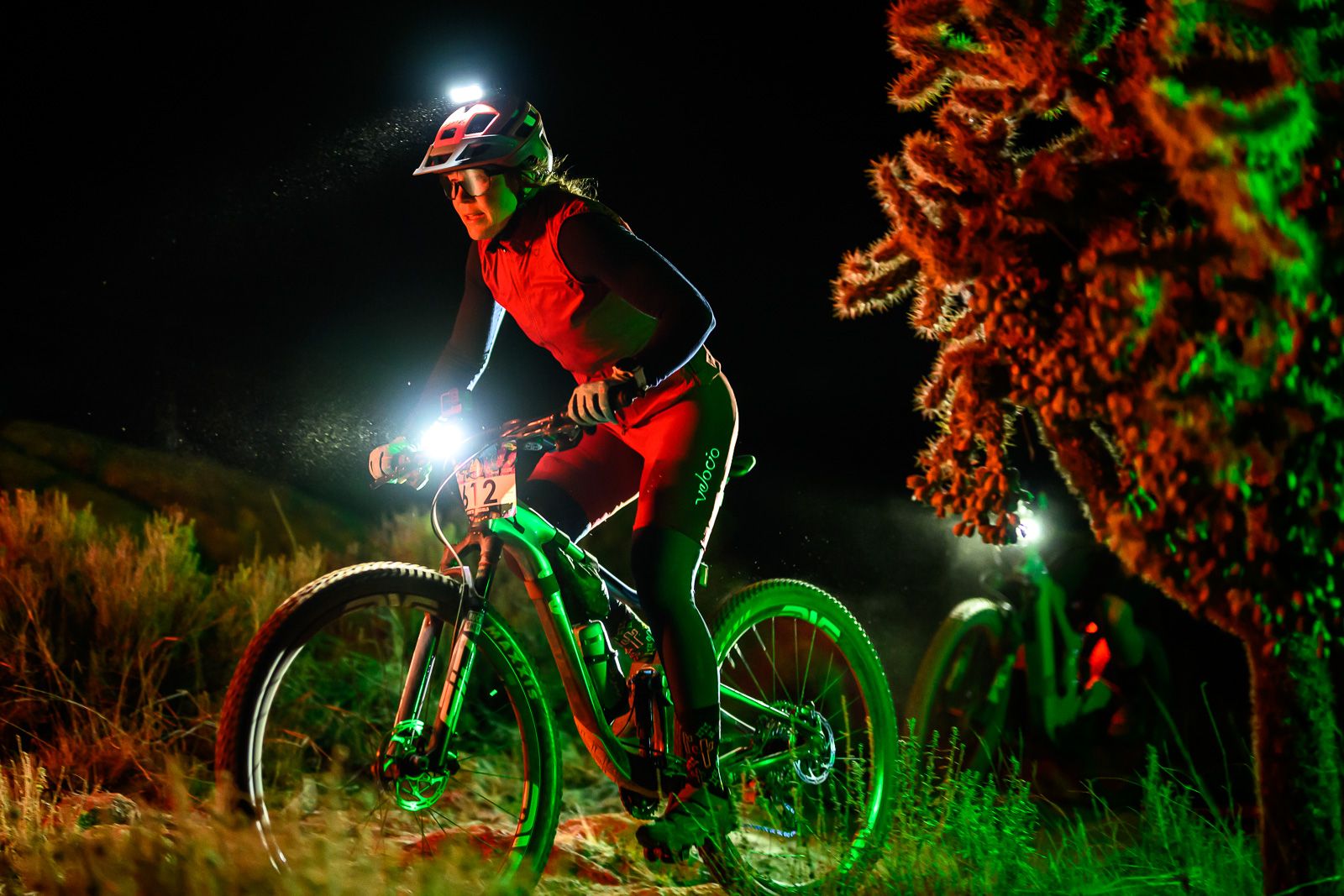 A cyclist wearing a helmet and headlamp rides a mountain bike at night, illuminated by green and red lights, with vegetation and another cyclist visible in the background.