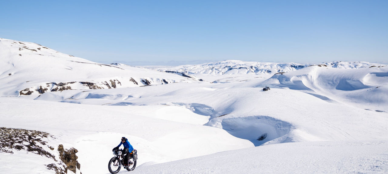 A person wearing winter gear rides a fat-tire bike across a snowy, mountainous landscape under a clear blue sky. Another cyclist is visible in the distance amid the snow-covered hills.