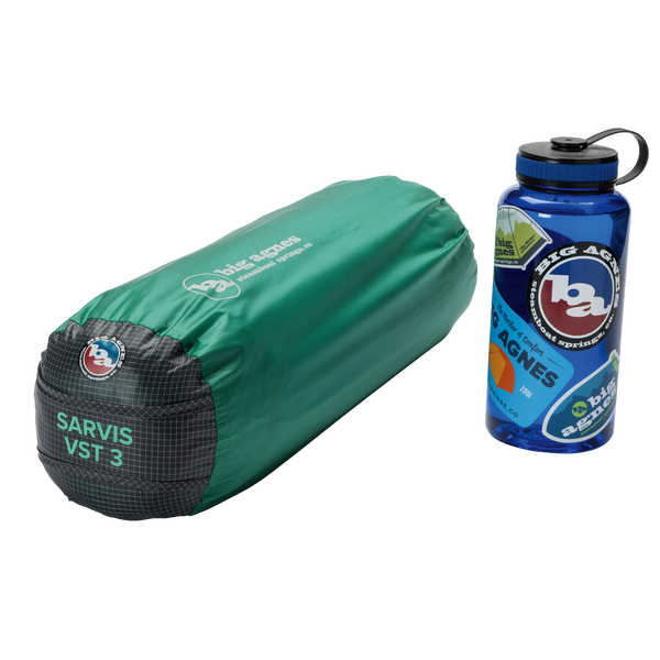 A green, cylindrical Sarvis VST 2 tent bag by Big Agnes Canada made with Hyperbead fabric rests beside a blue reusable water bottle with outdoor-themed stickers, both placed on a plain white background.