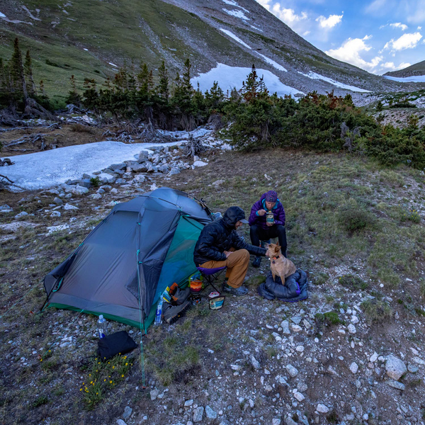 Two people relax near a Big Agnes Canada Sarvis VST 2 tent on a grassy mountain slope, sharing food with their dog. Snow patches, trees, and rocks surround them while camping gear and their ultralight shelter are scattered nearby.