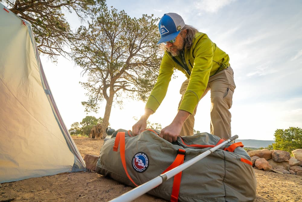 A person in outdoor clothing and a cap unpacks a large duffel bag with tent poles at a campsite, next to a pitched tent and a tree, under a bright sky.
