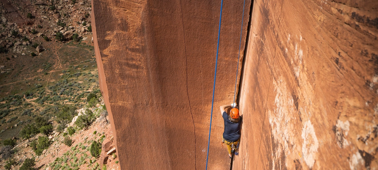 A rock climber wearing an orange helmet ascends a vertical, narrow crack in a tall sandstone cliff, secured by a blue rope, with a rugged landscape in the background.