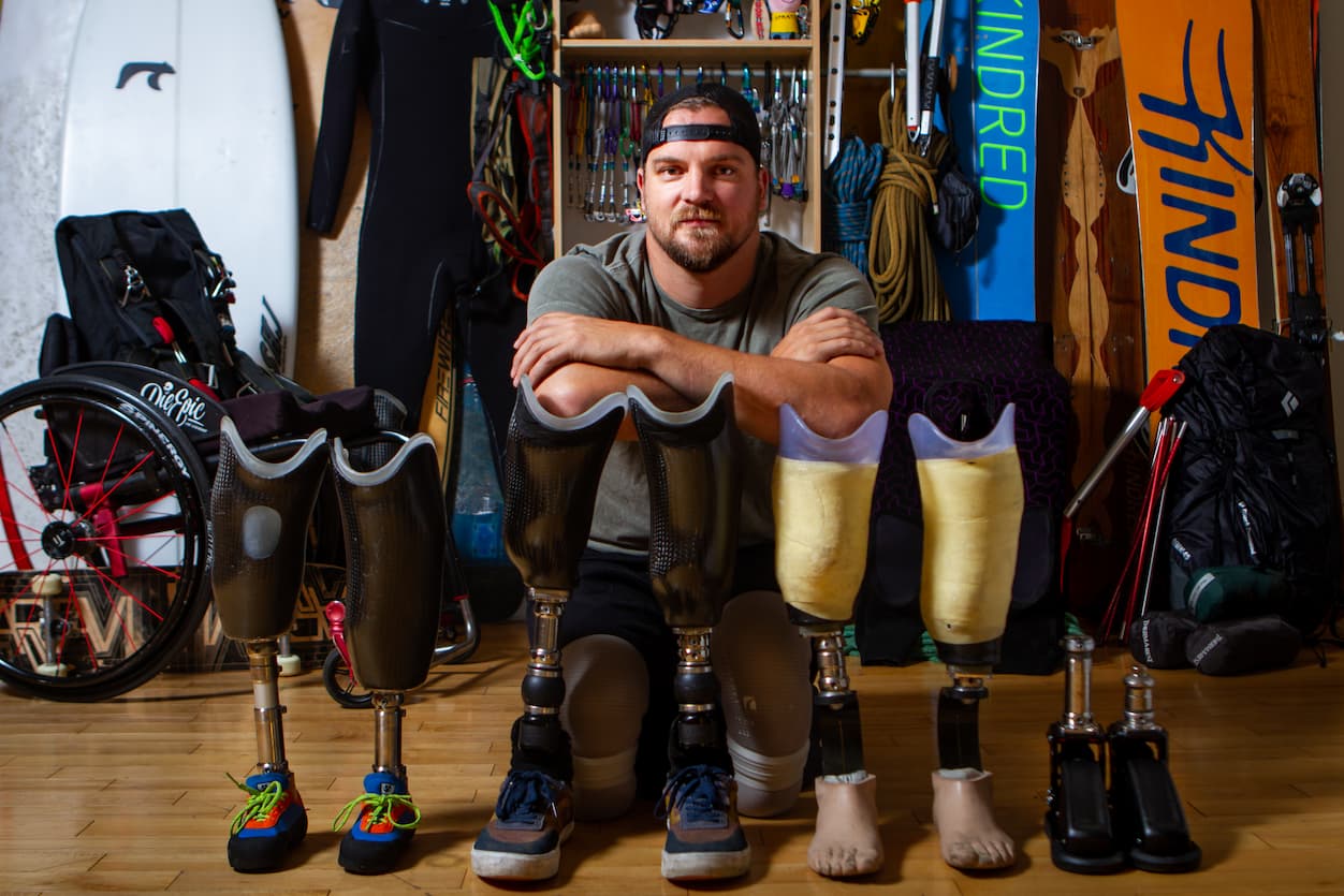A man with a beard and hat sits on the floor with his arms resting on his knees, surrounded by four pairs of prosthetic legs. Sports equipment, including surfboards and a wheelchair, are visible in the background.