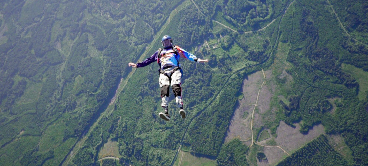 A person in a helmet and jumpsuit is skydiving over a landscape of green forests and fields, captured mid-air with arms and legs spread out.