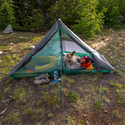 In a forest clearing, someone relaxes on an orange sleeping pad inside the Big Agnes Canada String Ridge VST 1.5 tent, reading a book. Shoes, a water bottle, and gear sit outside the ultralight shelter amid lush greenery.