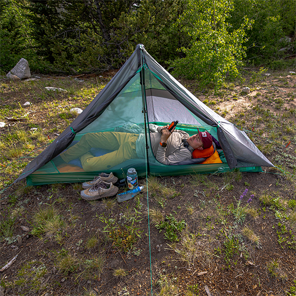 In a forest clearing, someone relaxes on an orange sleeping pad inside the Big Agnes Canada String Ridge VST 1.5 tent, reading a book. Shoes, a water bottle, and gear sit outside the ultralight shelter amid lush greenery.
