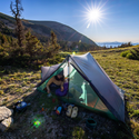 A person relaxes inside the Big Agnes Canada String Ridge VST 2.5 tent pitched on grassy terrain in the mountains, surrounded by camping gear under a clear blue sky with scenic hills in the background.