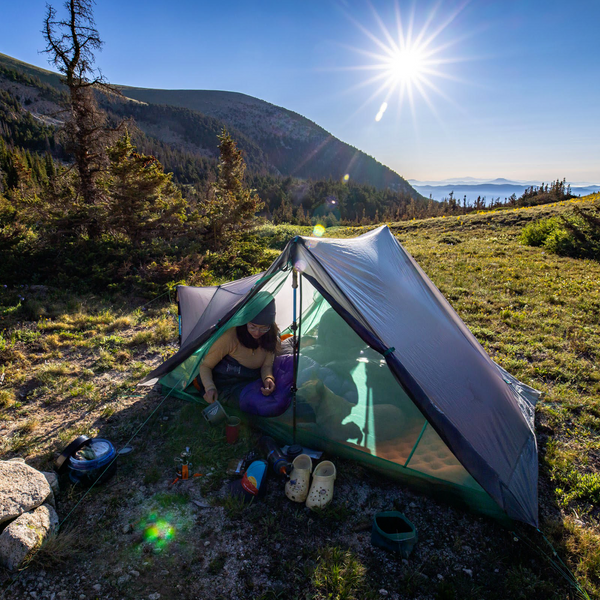 A person relaxes inside the Big Agnes Canada String Ridge VST 2.5 tent pitched on grassy terrain in the mountains, surrounded by camping gear under a clear blue sky with scenic hills in the background.